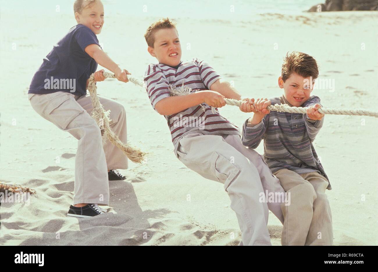 Strandszene, Portrait, blondes Maedchen und 2 Jungen im Alter von 10 -12 Jahren beim Tauziehen im Sand Stock Photo