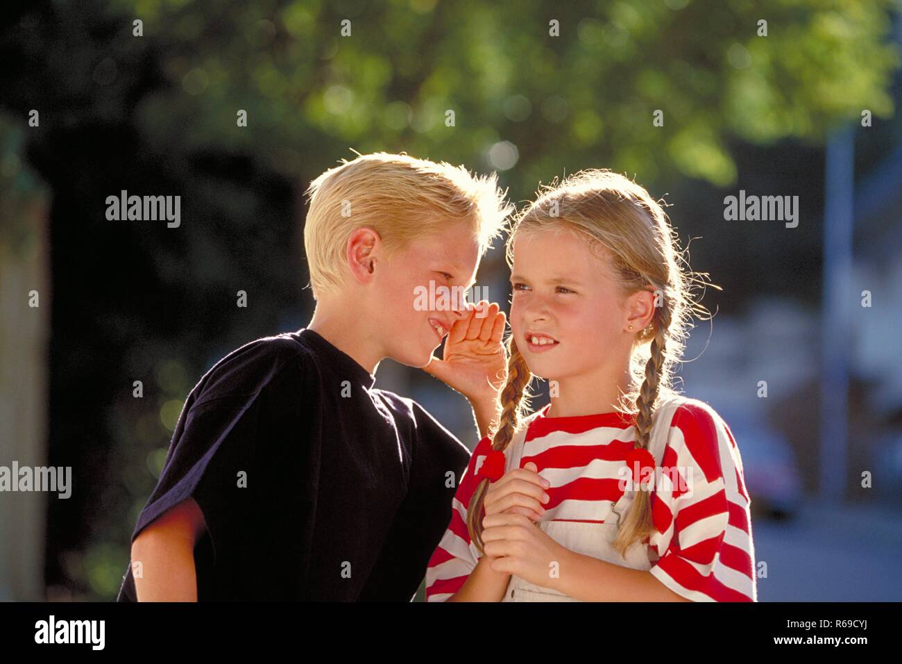 Outdoor, Halbfigur, blonder Junge erzaehlt blondem Maedchen mit Zoepfen , 8 Jahre alt, eine Neuigkeit Stock Photo