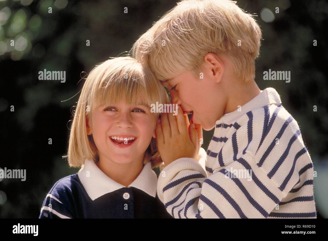 Outdoor, Portrait, blau-weiss gekleidetes blondes Kinderpaar, 5-6 Jahre, Junge fluestert dem  Maedchen etwas ins Ohr Stock Photo