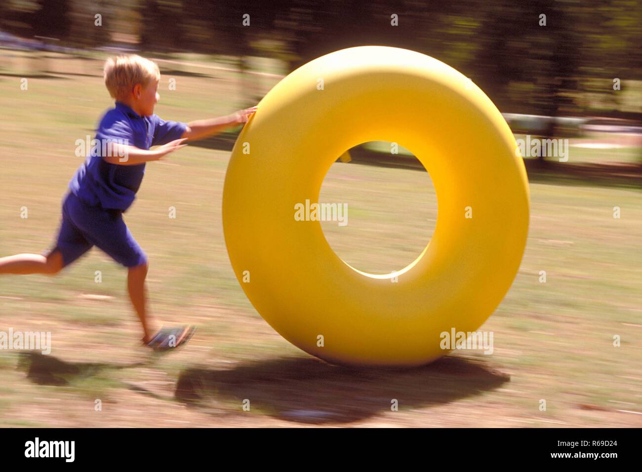 Outdoor, Parkszene, blau gekleideter blonder Jungen, 8 Jahre,  spielt mit einem grossen gelben Reifen auf einer Wiese Stock Photo