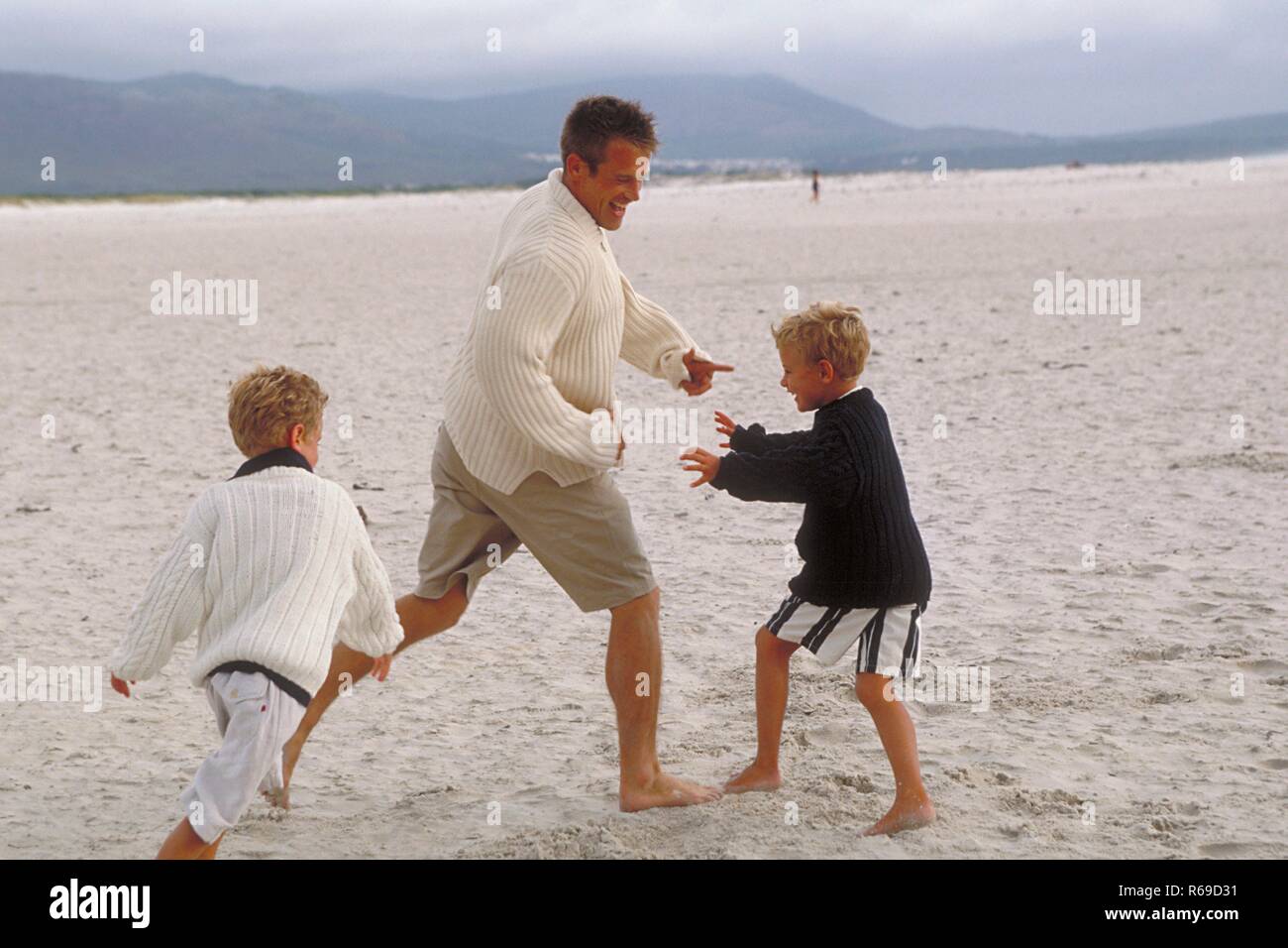 Halbfigur, Mann spielt mit seinen beiden 5-7 Jahre alten blonden Soehnen barfuss am Strand Stock Photo