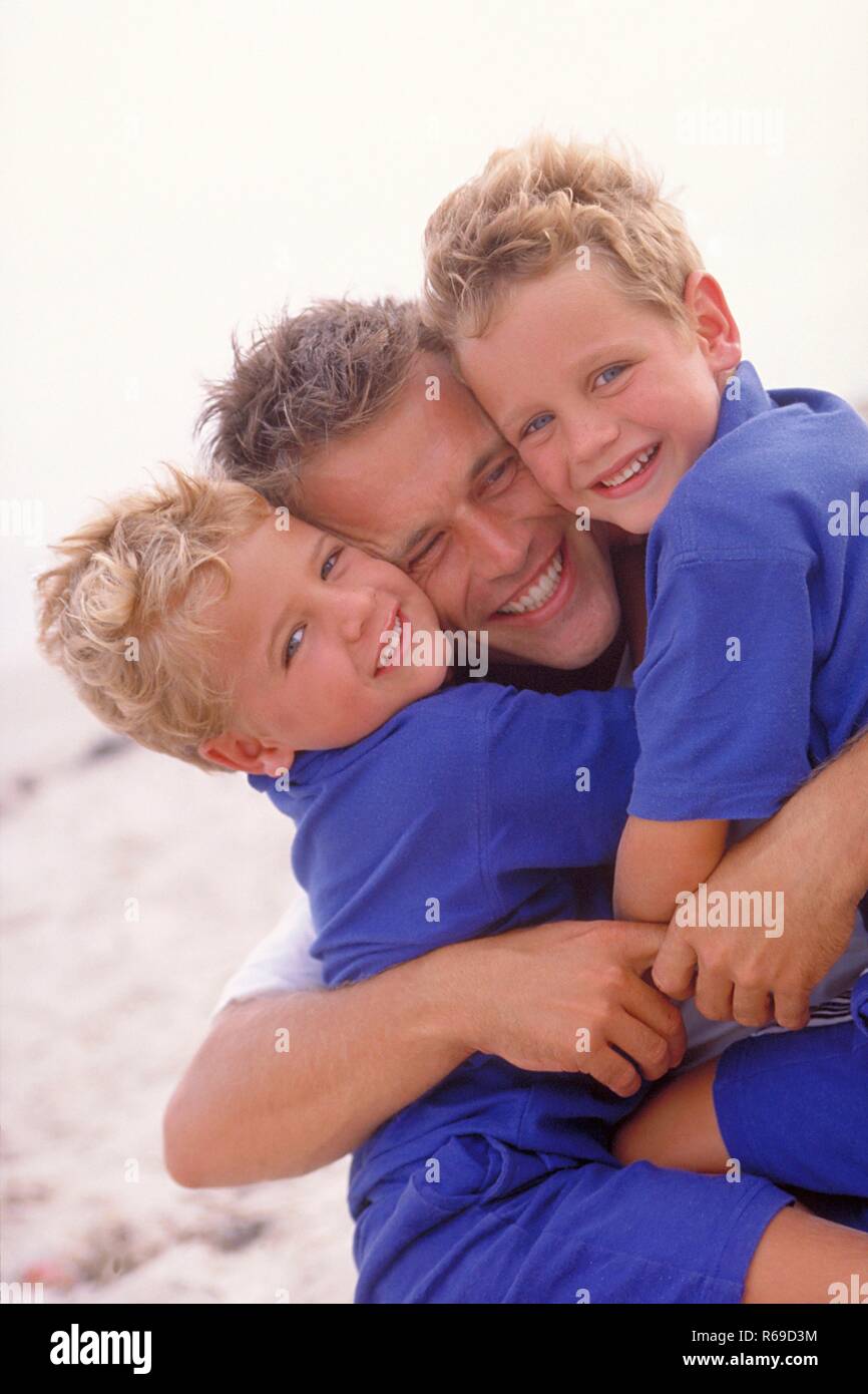 Portrait, Vater drueckt seine beiden 5-7 Jahre alten blonde Soehne, bekleidet mit blauen T-Shirts, am Strand  an sich Stock Photo