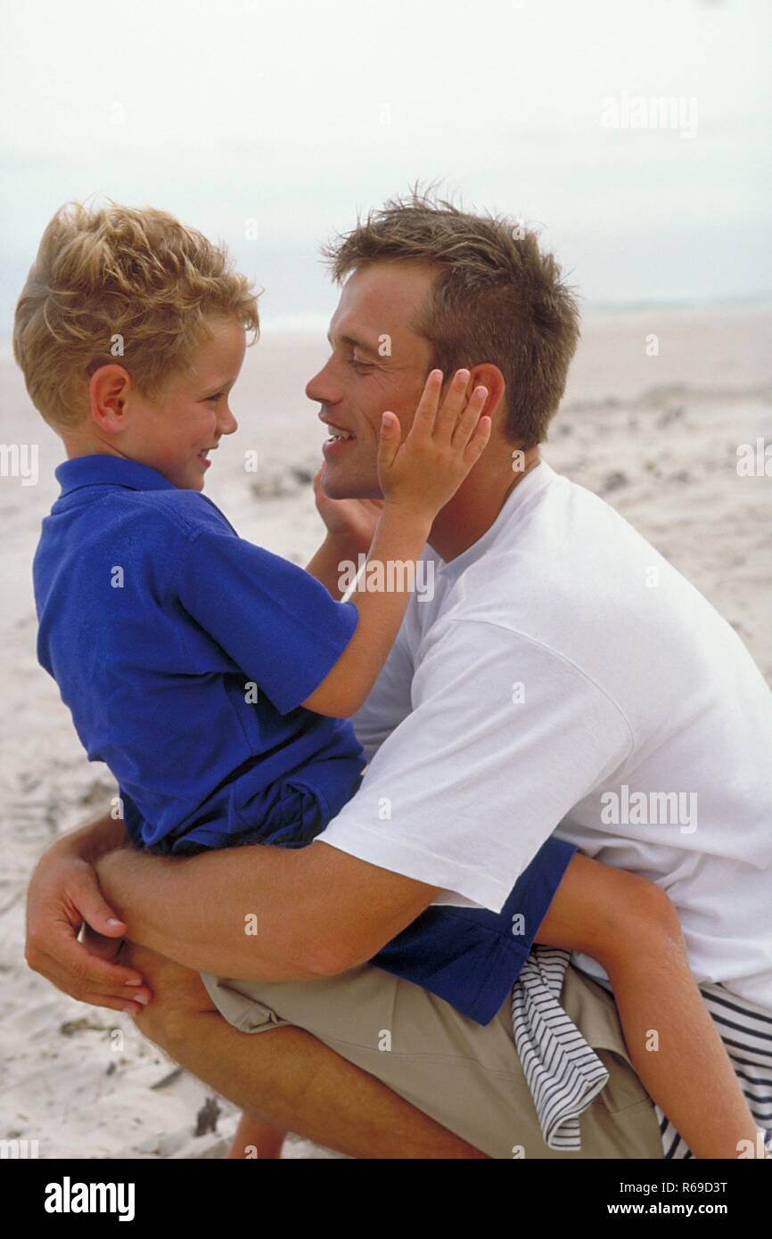 Strandszene, Vater sitzt mit seinem 6 Jahre alten blonden Sohn auf dem Schoss, bekleidet mit blauen T-Shirts, am Strand Stock Photo
