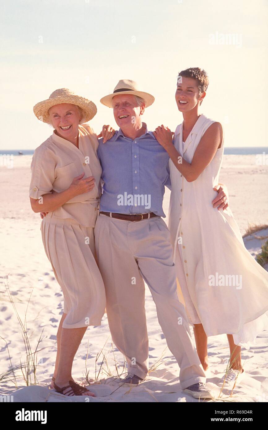 Strandszene, Ganzfigur, Portrait, Seniorenpaar bekleidet mit heller Sommerkleidung und Strohhut mit ihrer Tochter am Strand Stock Photo