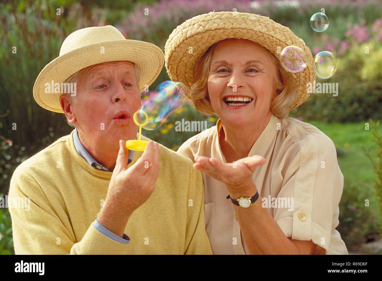 Outdoor, Portrait, Seniorenpaar bekleidet mit heller Kleidung und Strohhueten amuesieren sich mit Seifenblasen in einem Park Stock Photo