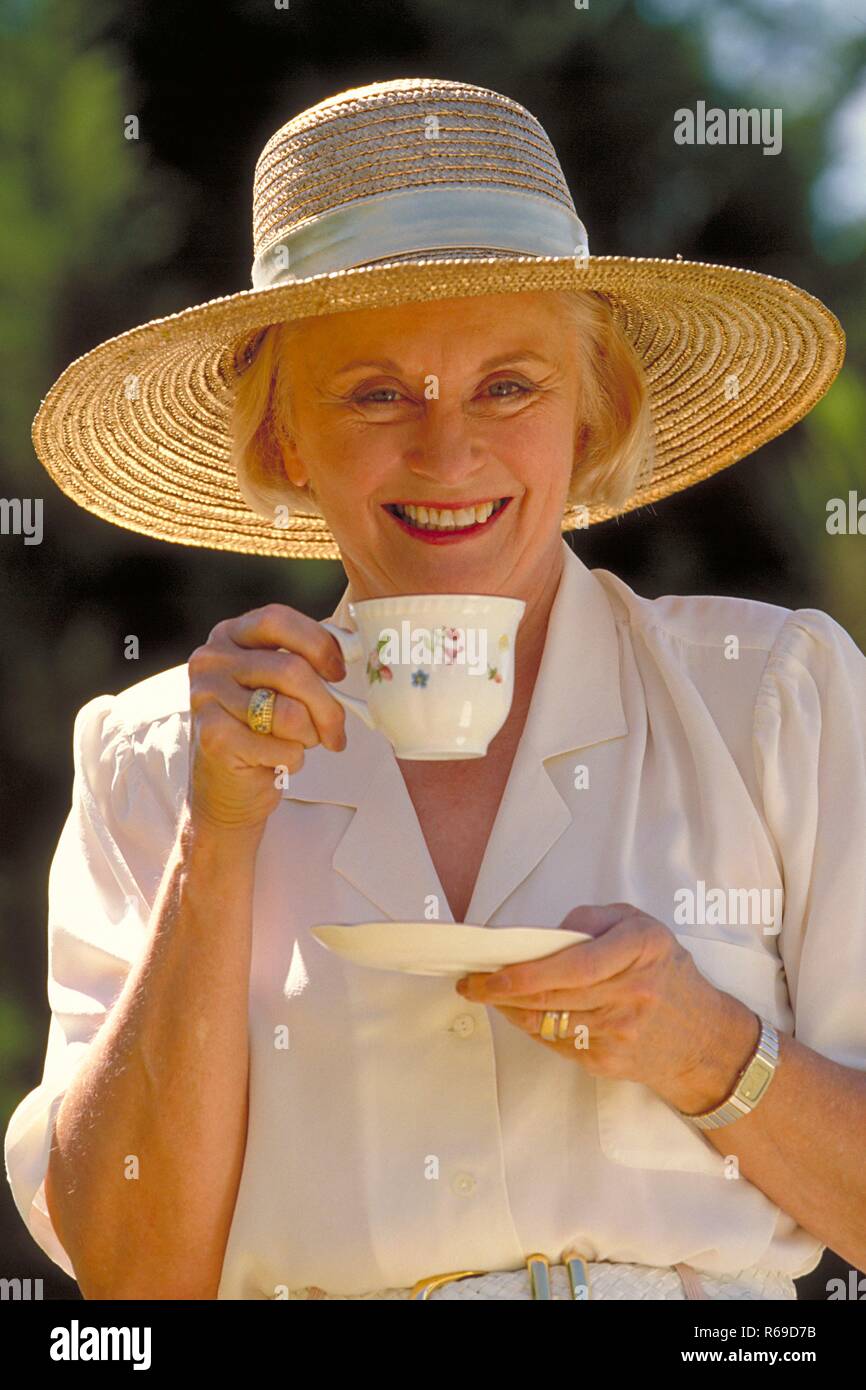 Outdoor, Portrait, Halbfigur, Frau Mitte 60 bekleidet mit weissem Hemd und Strohhut mit einer Tasse Kaffee in der Hand Stock Photo