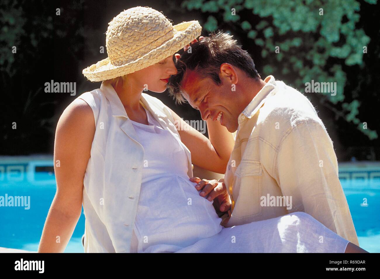 Outdoor, Profil, blonder Mann sitzt mit seiner schwangeren weiss gekleidete Frau mit Strohhut am Rand eines Schwimmbeckens Stock Photo