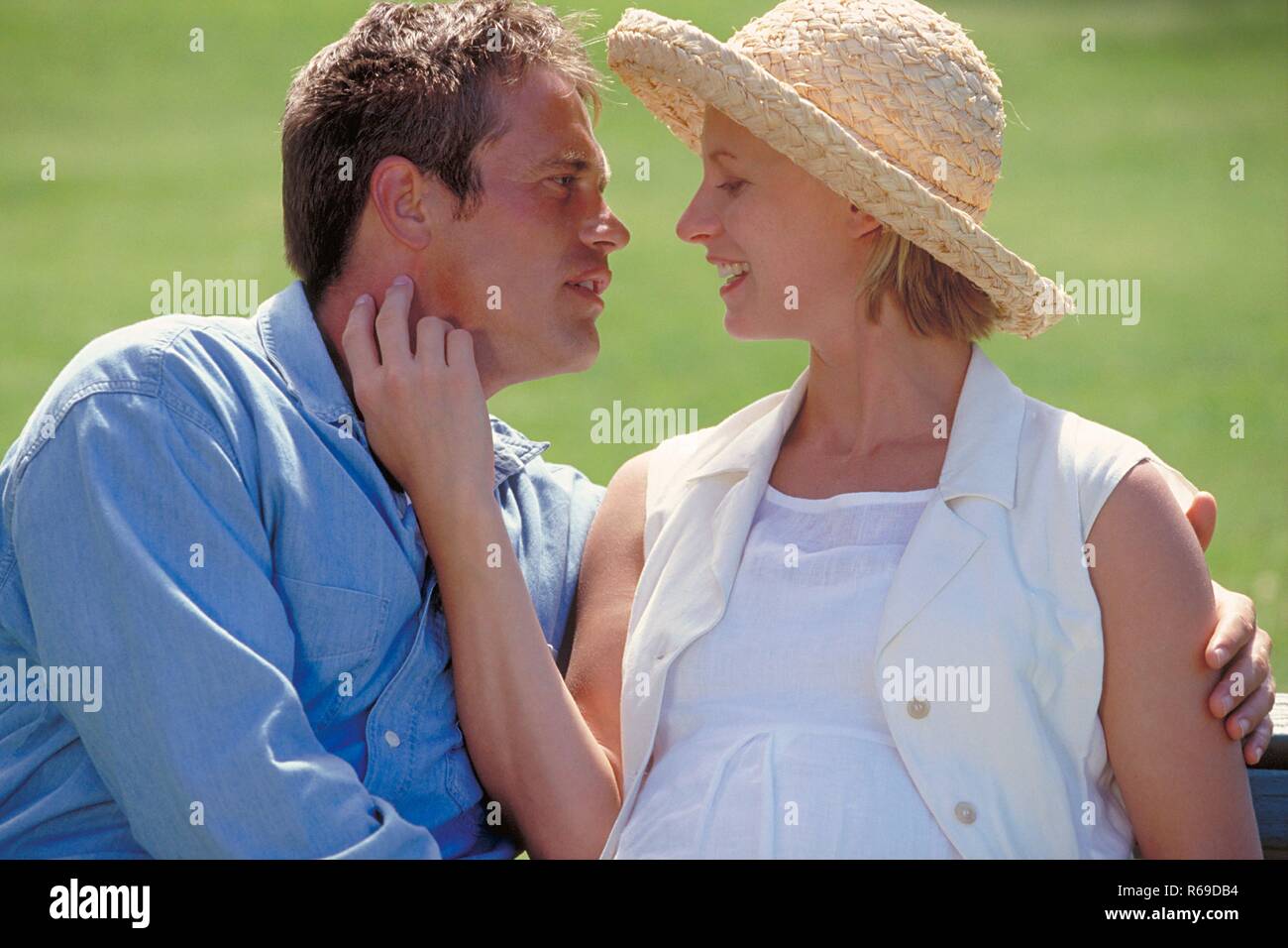 Outdoor, Parkszene, Profil, Mann im Jeanshemd und seine schwangere weiss gekleidete Frau mit Strohhut Stock Photo
