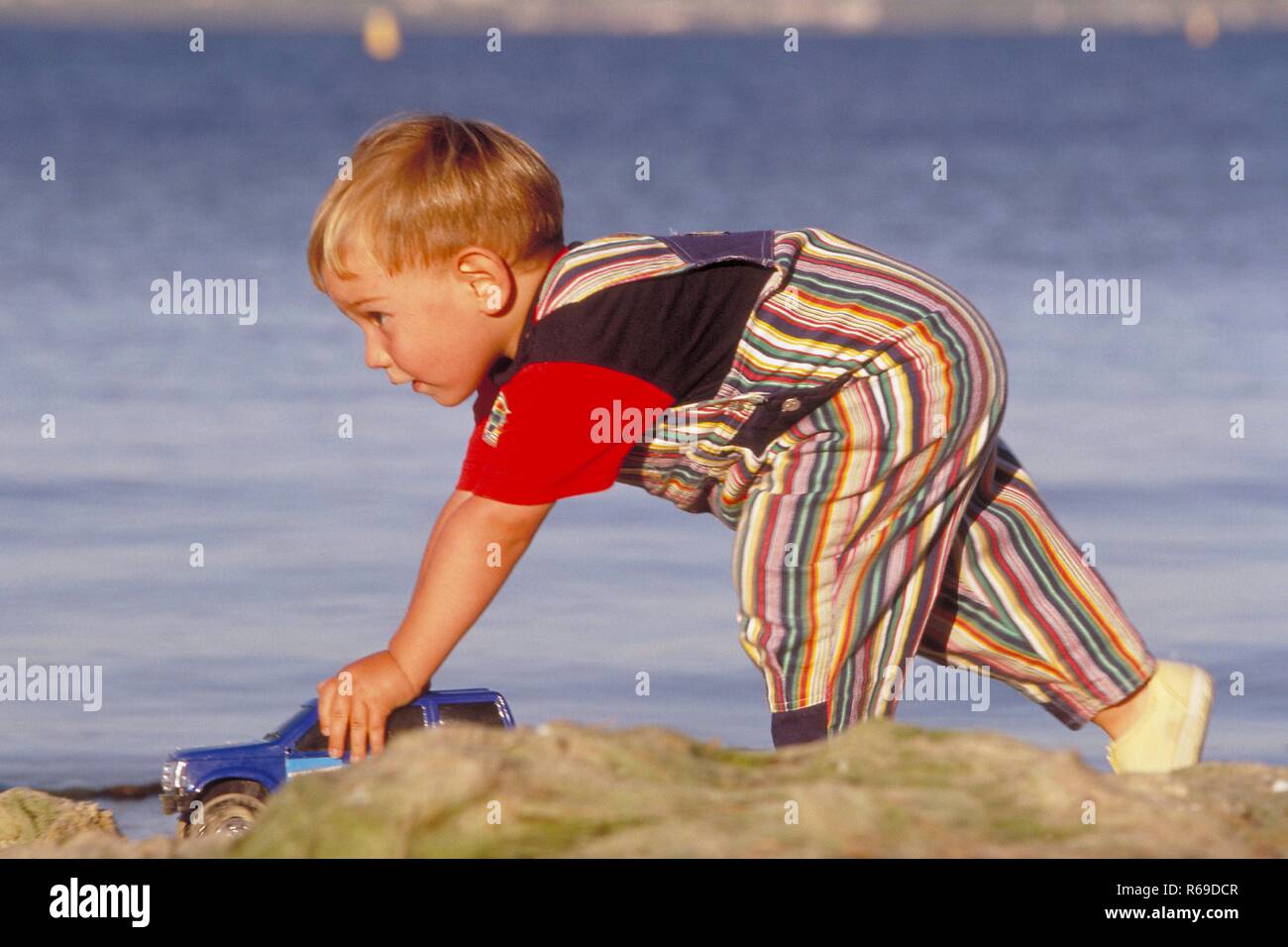 Ganzfigur, 3-jaehriger blonder Junge bekleidet mit gestreifter Latzhose spielt mit seinem Spielzeugjeep am Strand Stock Photo