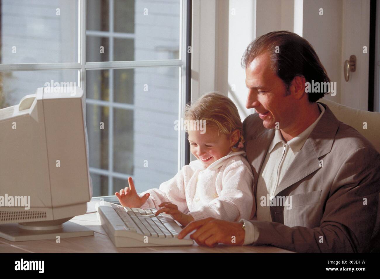 Innenraum, blondes Maedchen sitzt mit ihrem Vater am Tisch in der Naehe des Fensters vor dem Computer Stock Photo