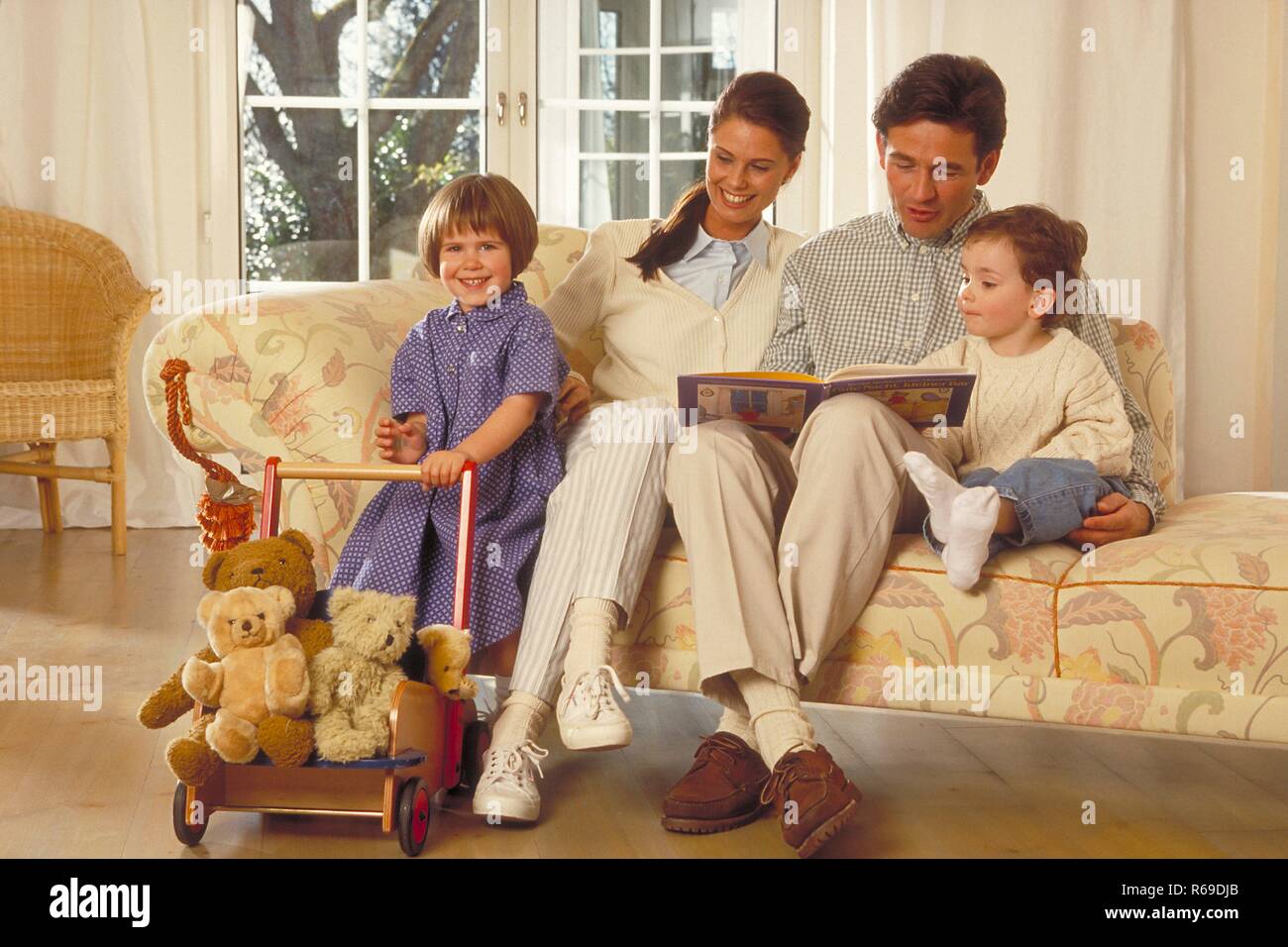 Innenraum, Gruppenbild, Familie, die Eltern sitzen mit ihren 2 Kindern auf dem Sofa, der Vater liest aus einem Bilderbuch vor Stock Photo