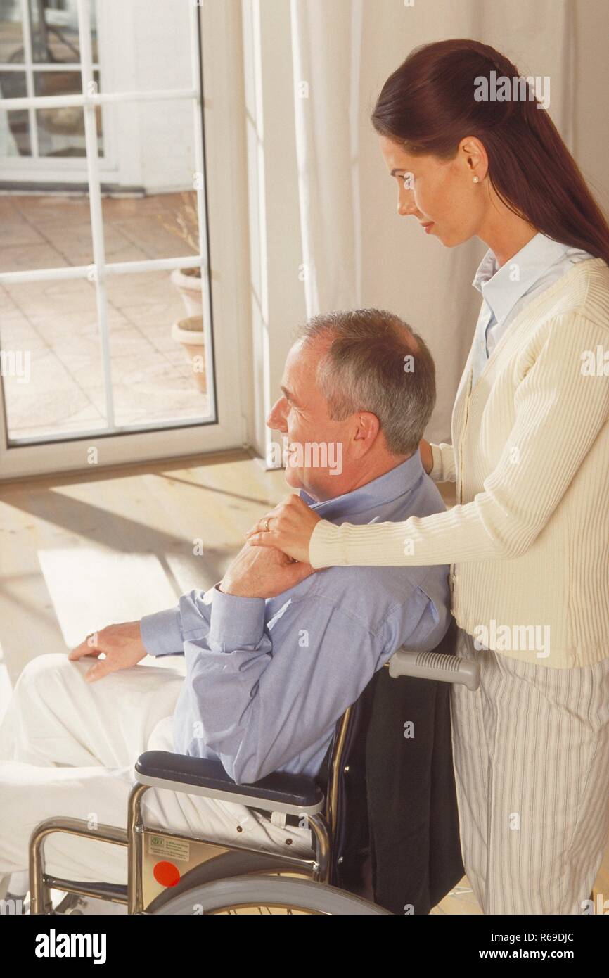 Portrait, Innenraum, Halbfigur, Frau mit langen braunen Haaren schiebt ihren im Rollstuhl sitzenden Vater vor eine Glastuer Stock Photo