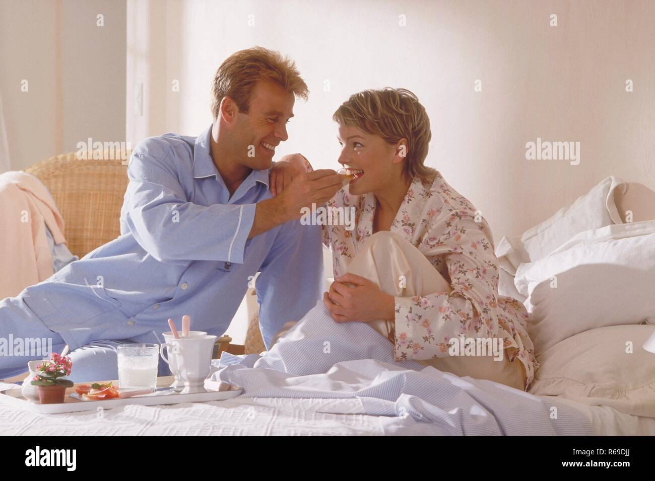 Portrait, Innenraum, Halbfigur, junges blondes Paar im Schlafanzug sitzt Sonntagmorgens beim Fruehstueck im Bett Stock Photo