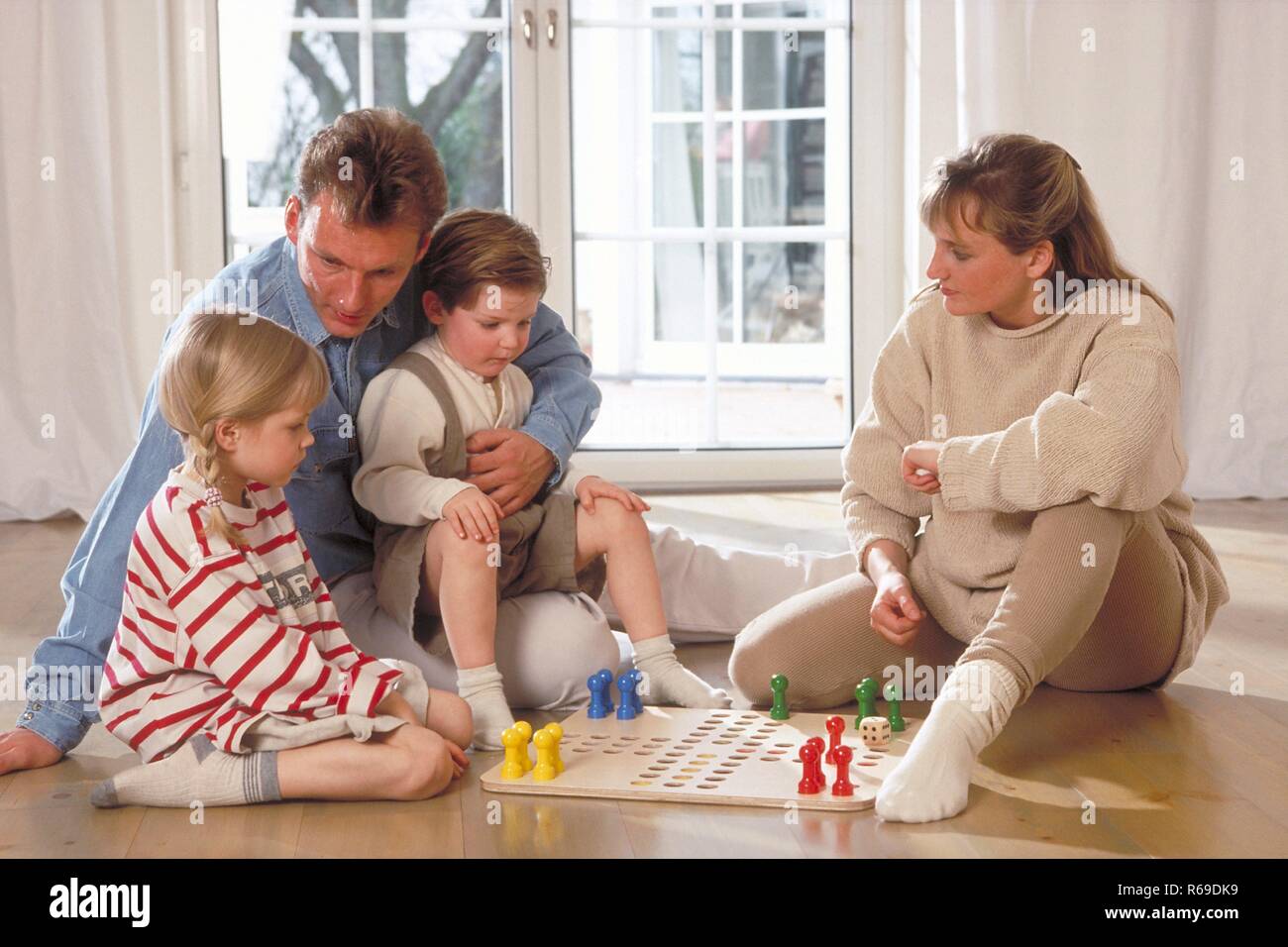 Innenraum, Ganzfigur, Familie mit 2 Kindern sitzt auf dem Fussboden vor dem Fenster und spielt eine Partie Mensch-aergere-dich-nicht Stock Photo