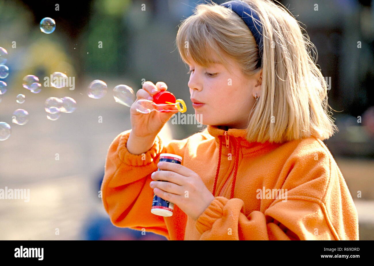 Portrait, Outdoor, blondes Maedchen mit orangem Sweater spielt mit Seifenblasen Stock Photo
