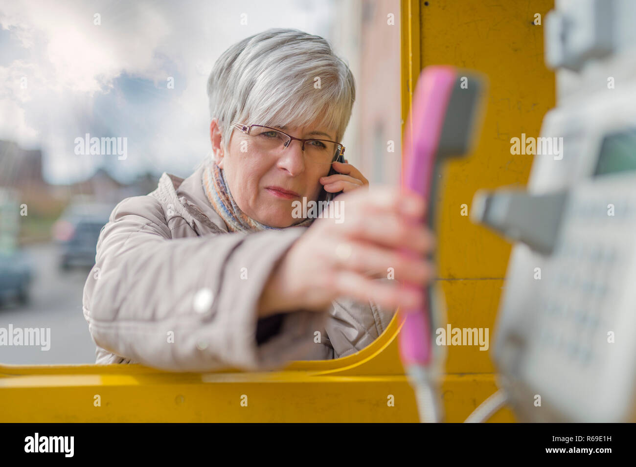 An Elderly Lady Stands On Smartphone In The Ear At A Phone Booth And Engages With His Hand Up The Phone. Stock Photo