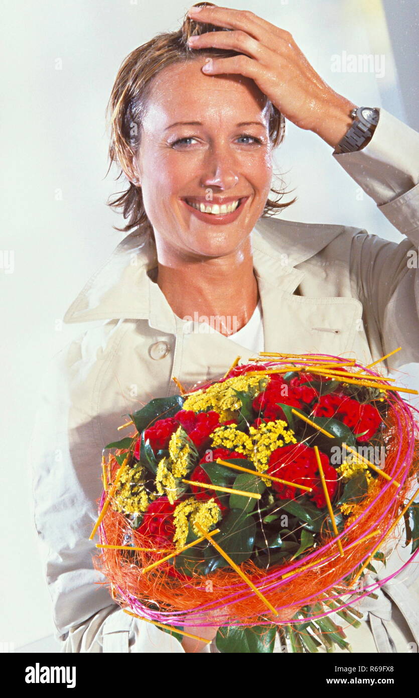 Portrait, Innenaufnahme, regennasse laechelnde 30 Jahre alte blonde Frau im hellen Trenchcaot mit einem bunten Blumenstrauss in der Hand Stock Photo