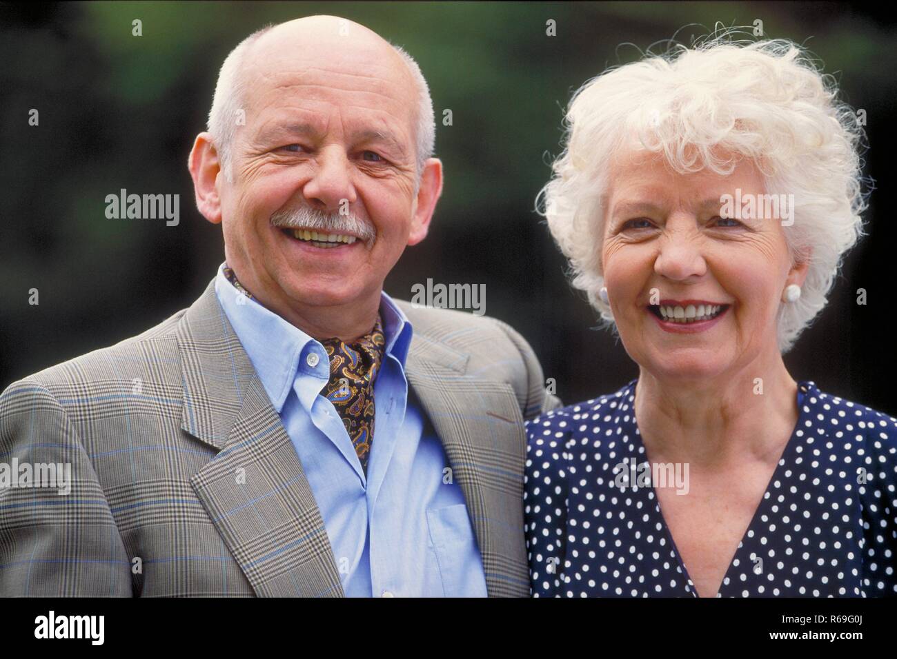 Portrait, Outdoor, laechelndes ca 75 Jahre altes Seniorenpaar, Mann mit Schnauzbart bekleidet mit gestreiftem Hemd, Jacket und Halstuch und weisshaarige Frau im blauen Kleid mit weissen Punkten Stock Photo