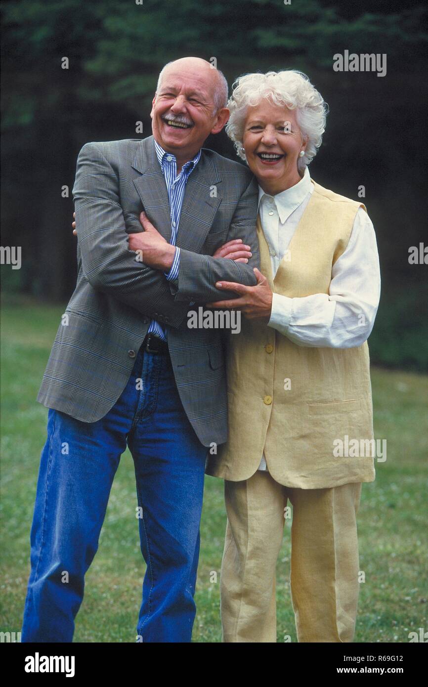 Portrait, Halbfigur, Outdoor, ca 75 Jahre altes Seniorenpaar  im Park, Mann mit Schnauzbart bekleidet mit Jeans und Jacket und weisshaarige Frau in gelber Hosenkombination Stock Photo