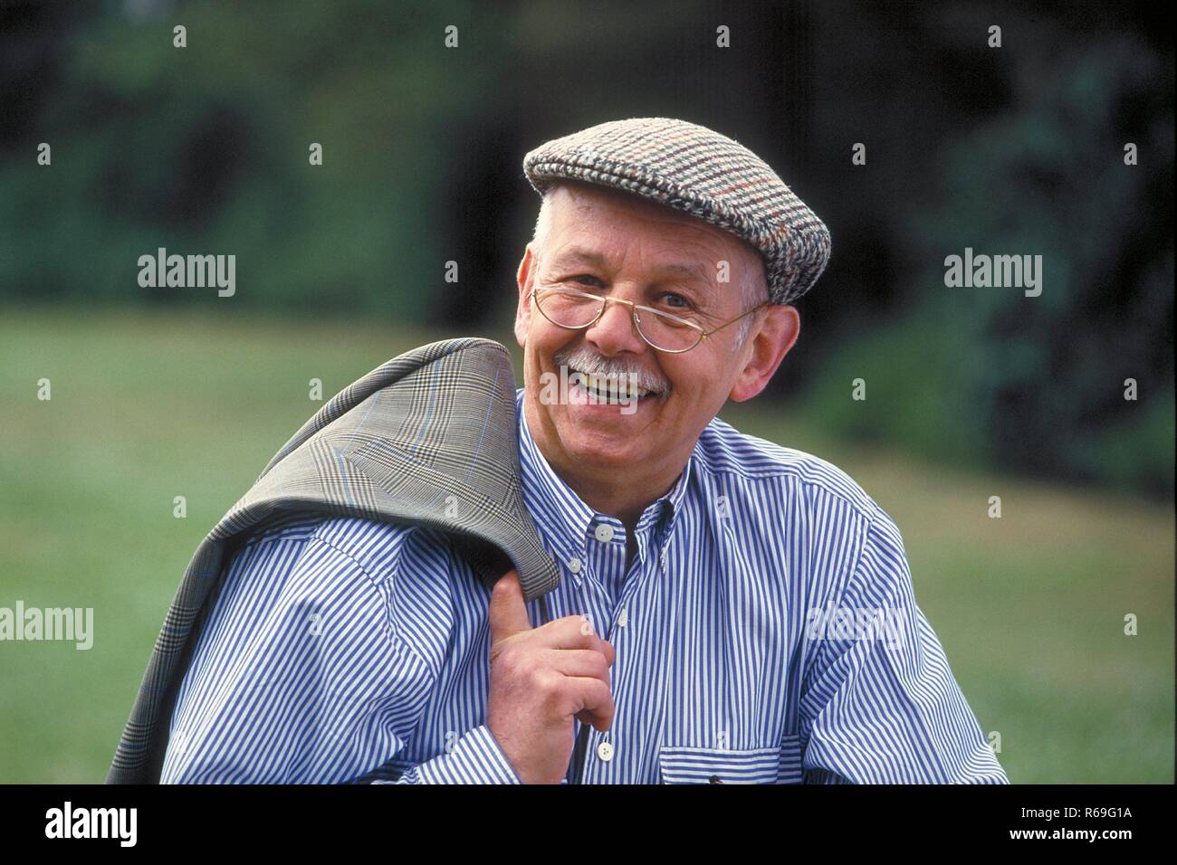Portrait, Outdoor, laechelnder Senior mit Schnauzbart, ca 75 Jahre alt, bekleidet mit blau-weiss gestreiftem Hemd, Schiebermuetze und Brille Stock Photo