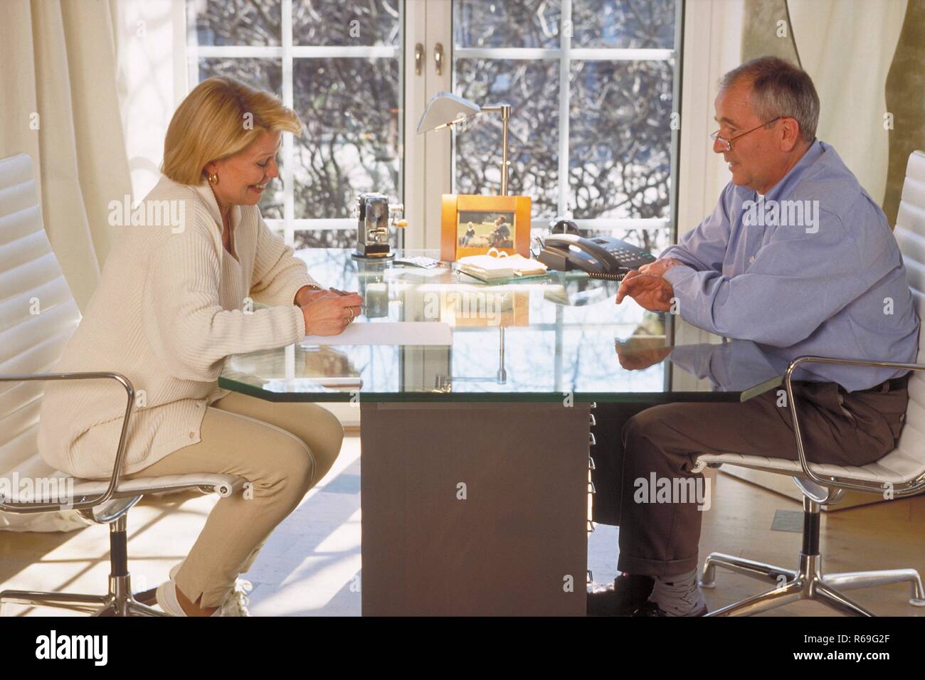 Innenraum, Ganzfigur, Profil, Seniorenpaar Mitte 60 sitzt sich am Glastisch vor dem Fenster gegenueber, die Frau macht Notizen Stock Photo