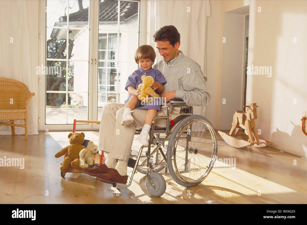 Portrait, Innenraum, Ganzfigur,  Mann sitzt mit seiner  4-jaehrigen Tochter und ihrem Teddy auf dem Schoss im Rollstuhl vor einer Glastuer Stock Photo
