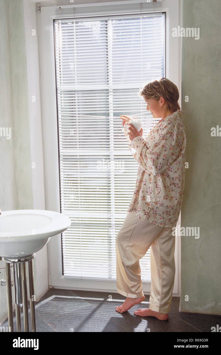 Portrait, Innenraum, Ganzfigur, blonde Frau im Schlafanzug steht barfuss mit einer Tasse Kaffee in der Hand im Badezimmer an einer Glastuer und schaut nach draussen Stock Photo