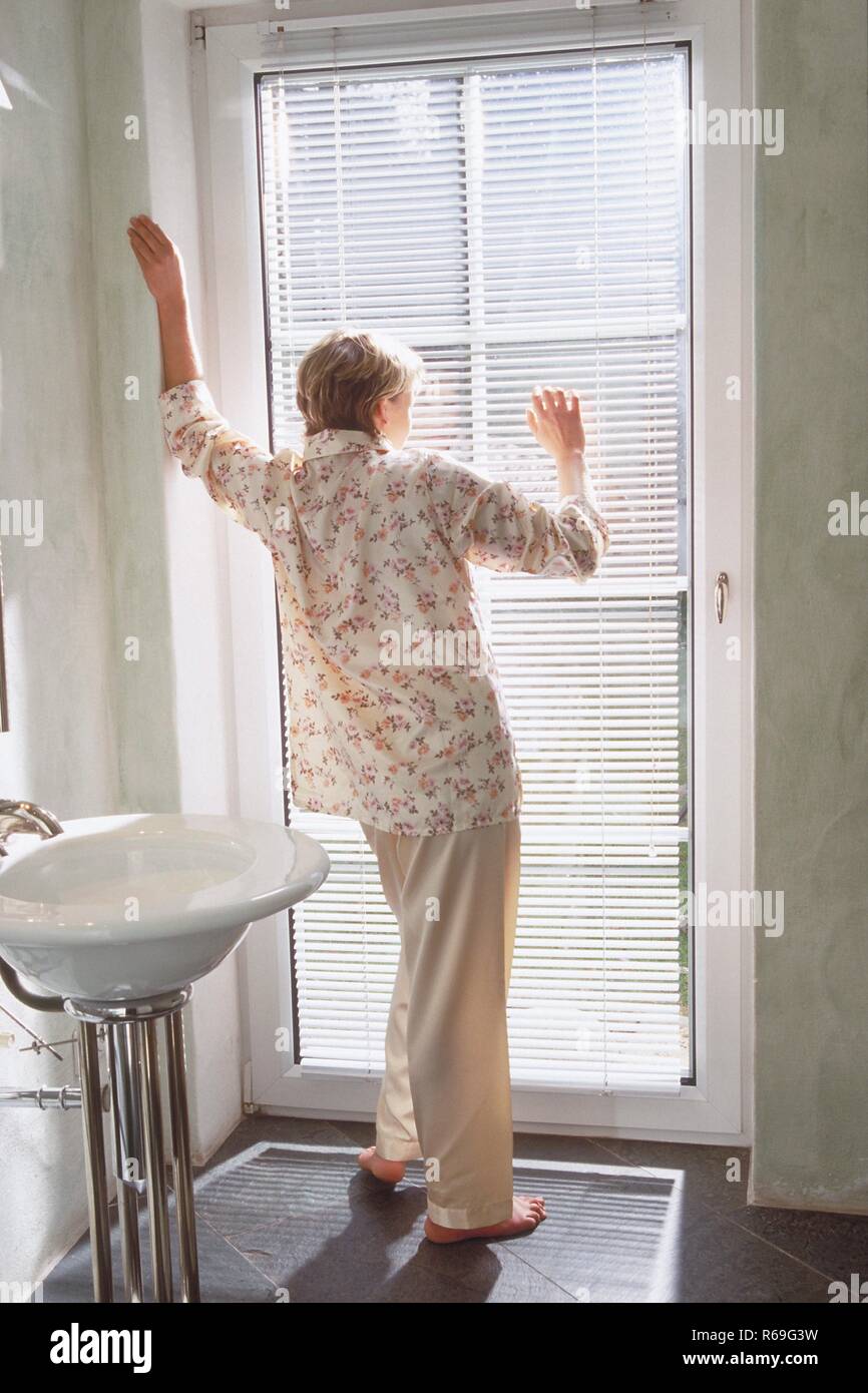 Portrait, Innenraum, Ganzfigur, blonde Frau im Schlafanzug steht barfuss im Badezimmer an einer Glastuer und schaut nach draussen Stock Photo