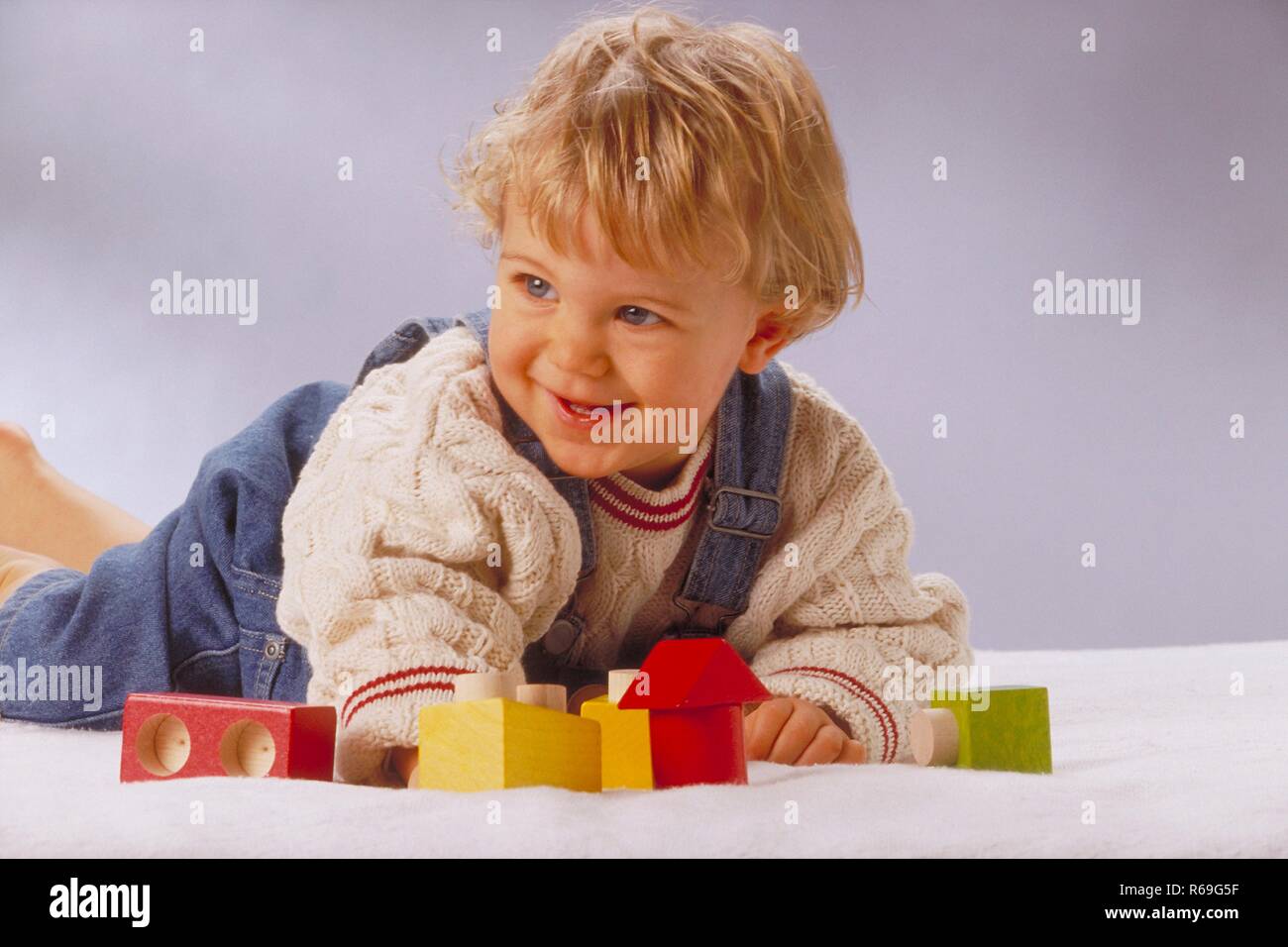 Portrait,  Innenraum, 1-jaehriger blonder Junge bekleidet mit Jeanslatzhose und weissem Pullover liegt auf dem Boden und spielt mit bunten Baukloetzen Stock Photo