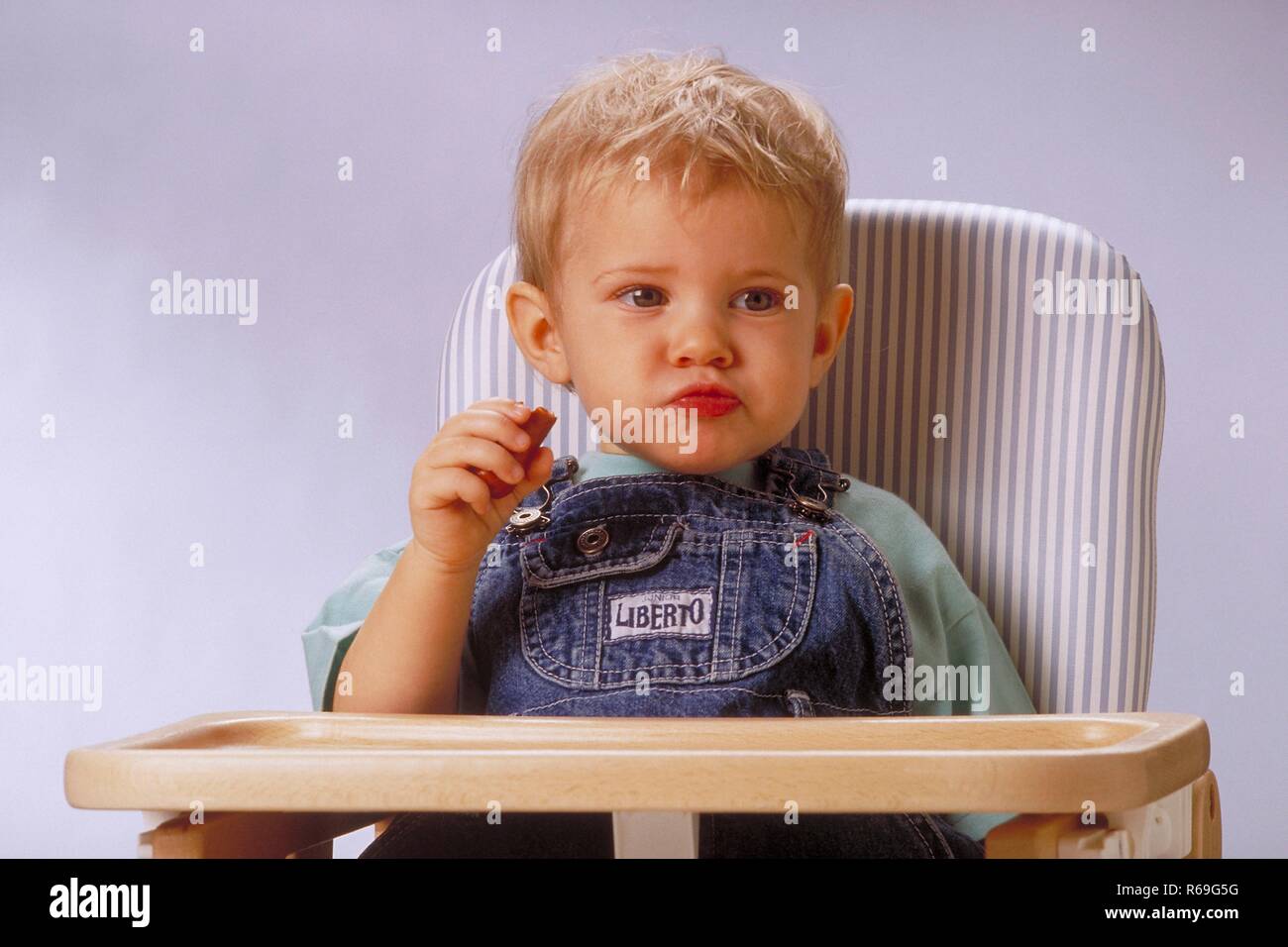 Portrait,  Innenraum, ca. 1-jaehriger blonder Junge bekleidet mit Jeanslatzhose sitzt mit einem Wuerstchen in der Hand in einem Hochstuhl Stock Photo