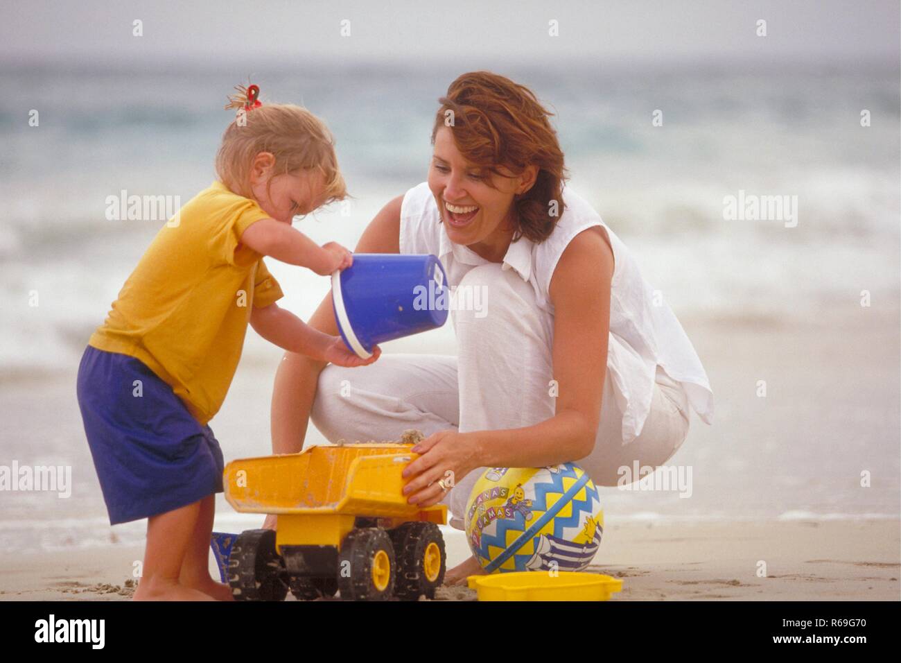 Ganzfigur, Outdoor, weiss gekleidete Frau spielt mit ihrer 2-jaehrigen blonden Tochter mit einem blauen Eimer und gelbem Spielzeuglaster am Strand Stock Photo