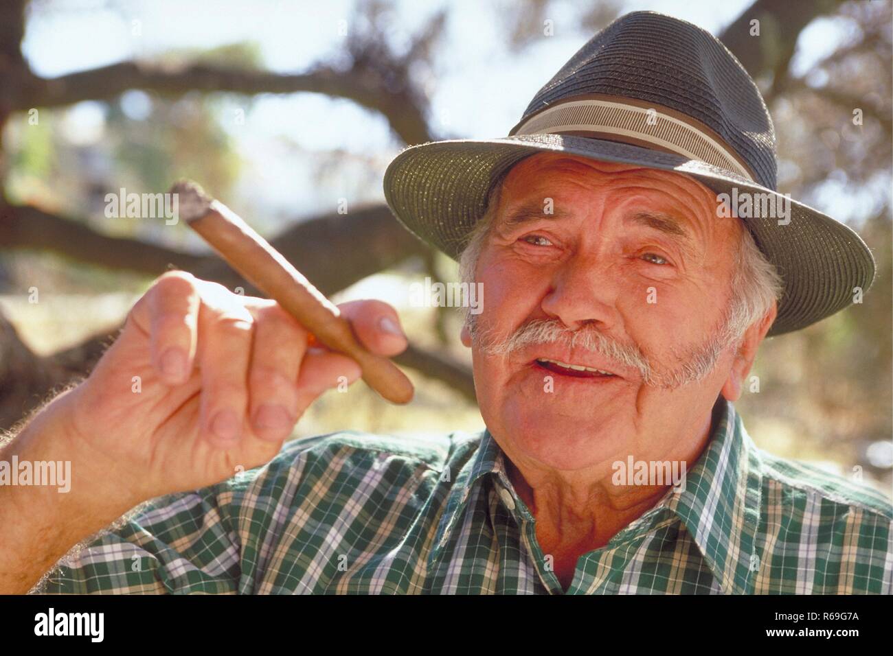 Portrait, Outdoor, Mann mit Oberlippenbart, ca. 65 Jahre alt, bekleidet mit gruen-weiss kariertem Hemd und Hut mit einer Zigarre in der Hand unter einem Baum Stock Photo