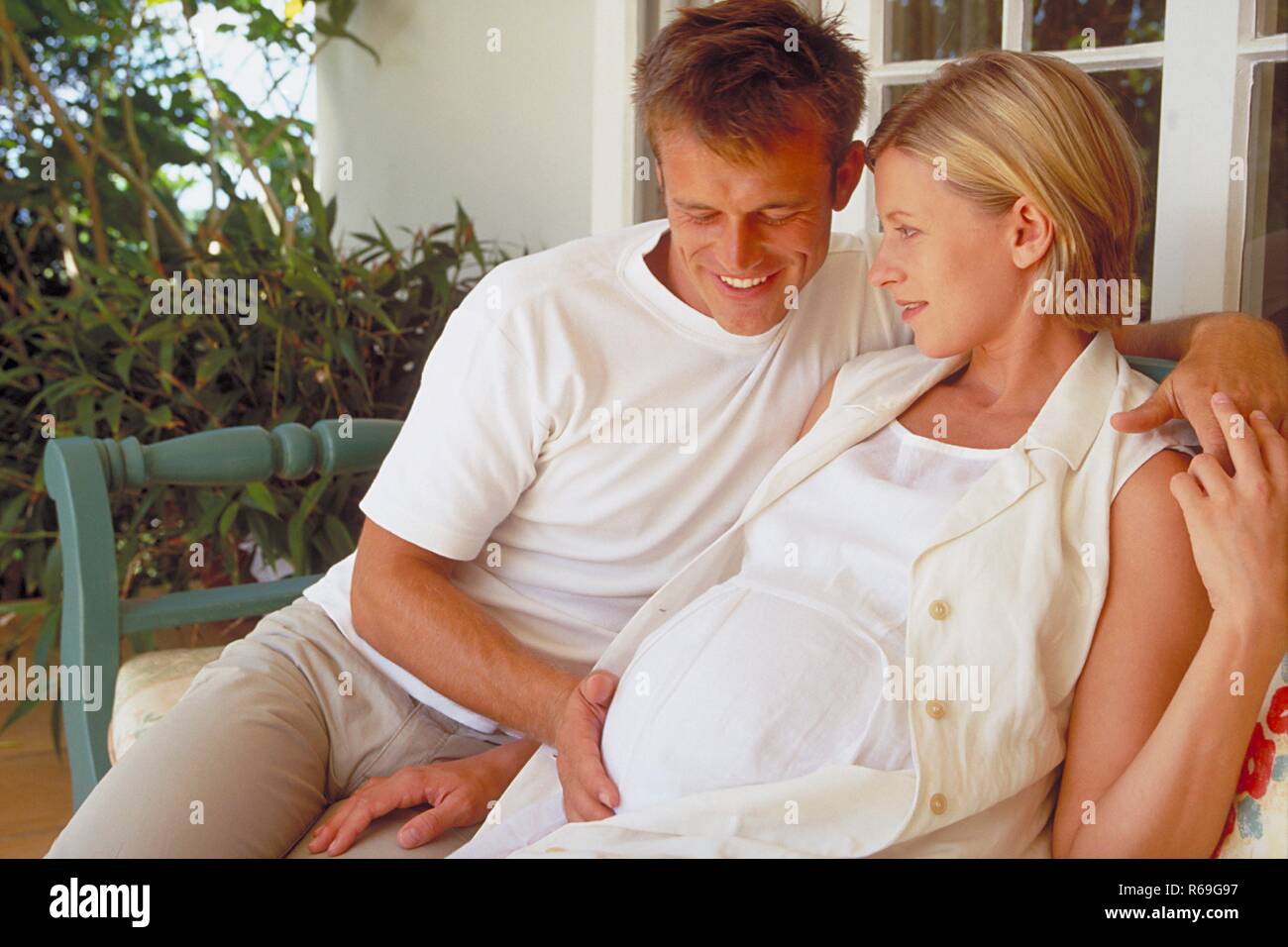 Outdoor, hell gekleideter Mann sitzt mit seiner schwangeren weiss gekleideten Frau auf einer Bank auf der Terrasse und streichelt ihren Bauch Stock Photo