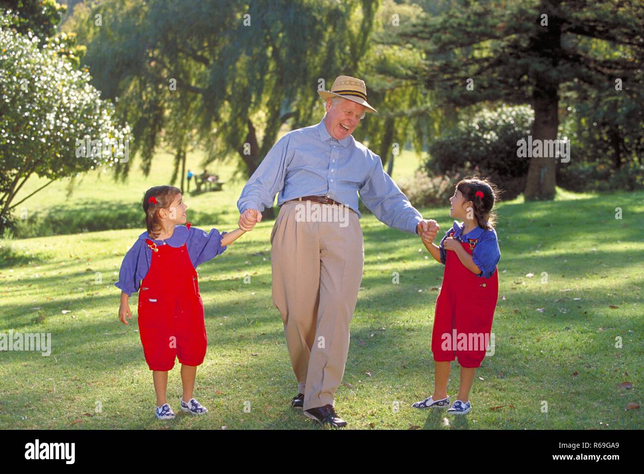 Outdoor, Parkszene, Grossvater geht mit zwei 6 Jahre alte Zwillingsmaedchen an der Hand, bekleidet mit roten Latzhosen und blauen Blusen, lachend ueber eine Wiese Stock Photo