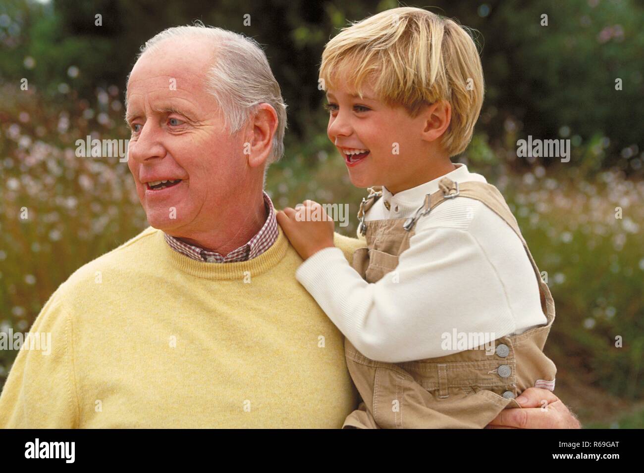 Outdoor, blonder 6 Jahre alter Junge bekleidet mit beiger Latzhose auf dem Arm seines Grossvaters bekleidet mit gelbem Pullover Stock Photo