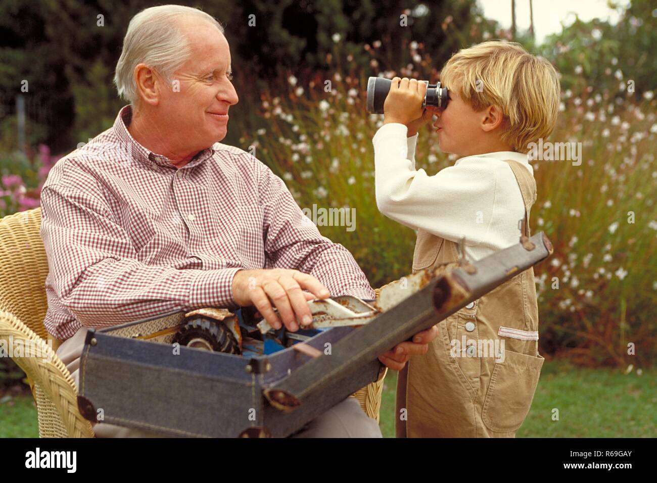 Outdoor, blonder 6 Jahre alter Junge bekleidet mit beiger Latzhose steht mit einem Fernglas neben seinem Grossvater, der mit einem Koffer Spielsachen auf den Beinen auf einem Korbsessel im Garten sitzt Stock Photo