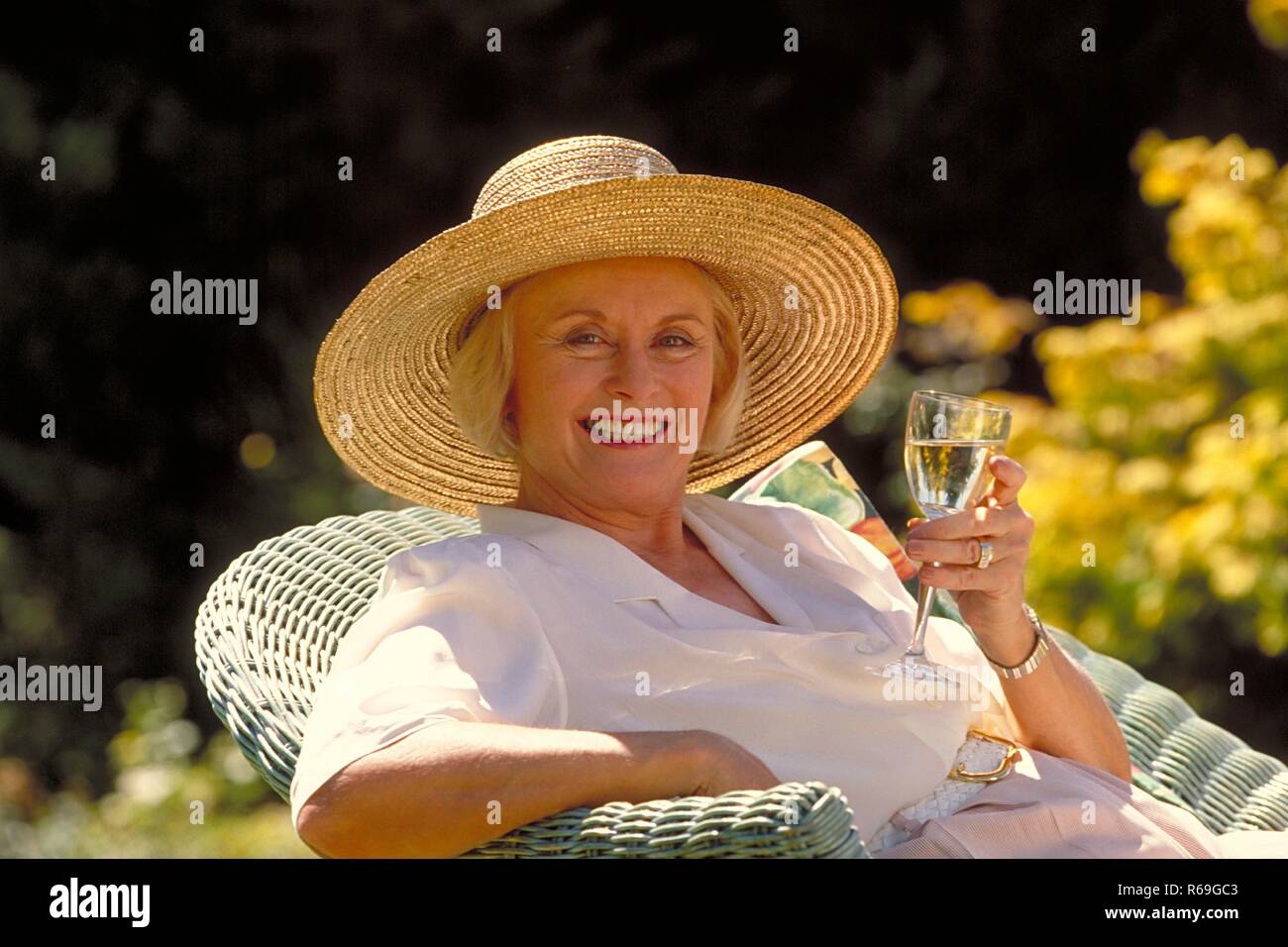 Outdoor, Portrait, blonde Frau Mitte 60 bekleidet mit weisser Bluse und Strohhut sitzt mit einem Glas Sekt auf einem Korbsessel im Garten Stock Photo