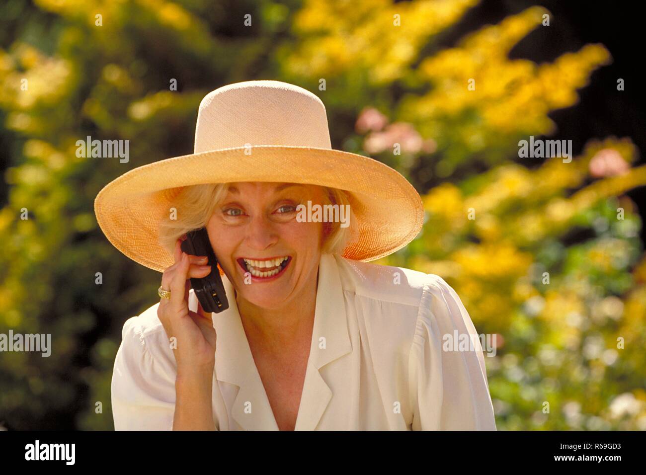 Outdoor, Portrait, Halbfigur, Frau Mitte 60 bekleidet mit weissem Hemd und  Strohhut schneidet telefoniert im Garten mit einem Handy Stock Photo