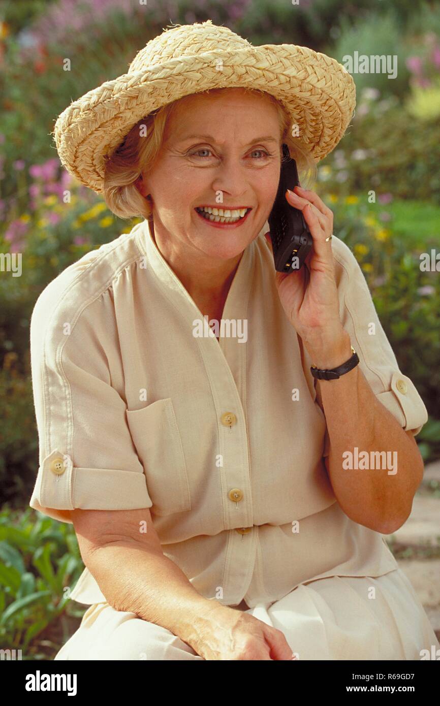 Outdoor, Portrait, Halbfigur, Frau Mitte 60 bekleidet mit beiger Bluse, Rock und Strohhut sitzt im Garten und telefoniert mit einem Handy Stock Photo