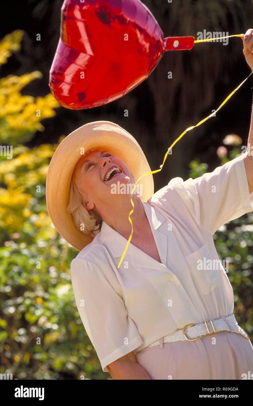 Outdoor, Portrait, Halbfigur, Frau Mitte 60 bekleidet mit heller  Hose,weissem Hemd und Strohhut spielt mit einem roten herzfoermigen Luftballon Stock Photo