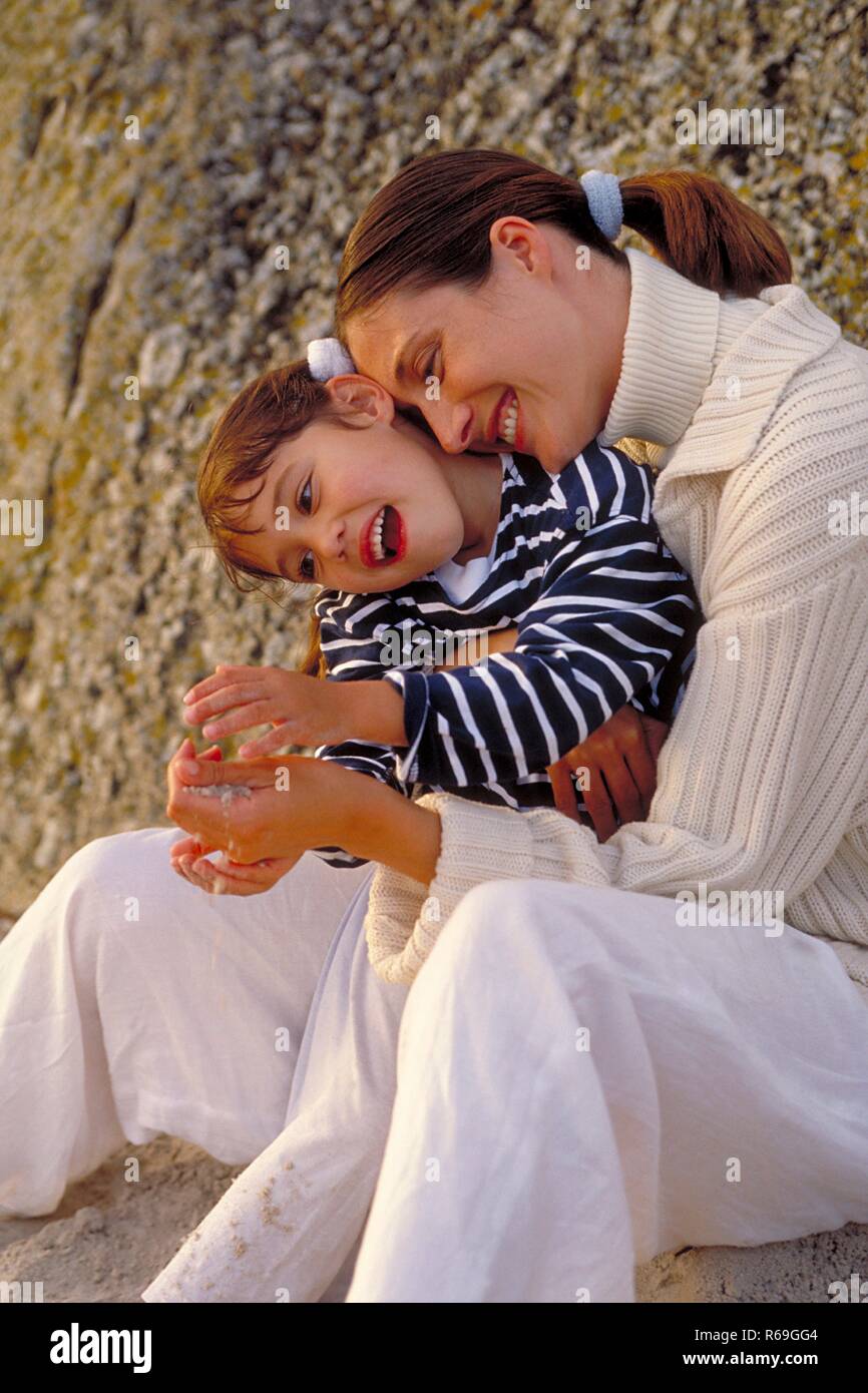 Strandszene, Halbfigur, bruenette Mutter bekleidet mit hellem Pulli und Tochter, 8 Jahre alt, bekleidet mit gestreiftem Pulli sitzen umschlungen vor einem Felsen im Sand Stock Photo