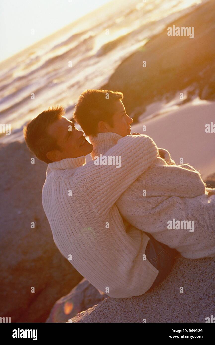 Strandszene, Halbfigur, Mann und Frau mit kurzen braunen Haaren bekleidet mit naturfarbenen Wollpullovern und Jeans sitzen bei Sonnenuntergang auf einem Felsen am Strand Stock Photo