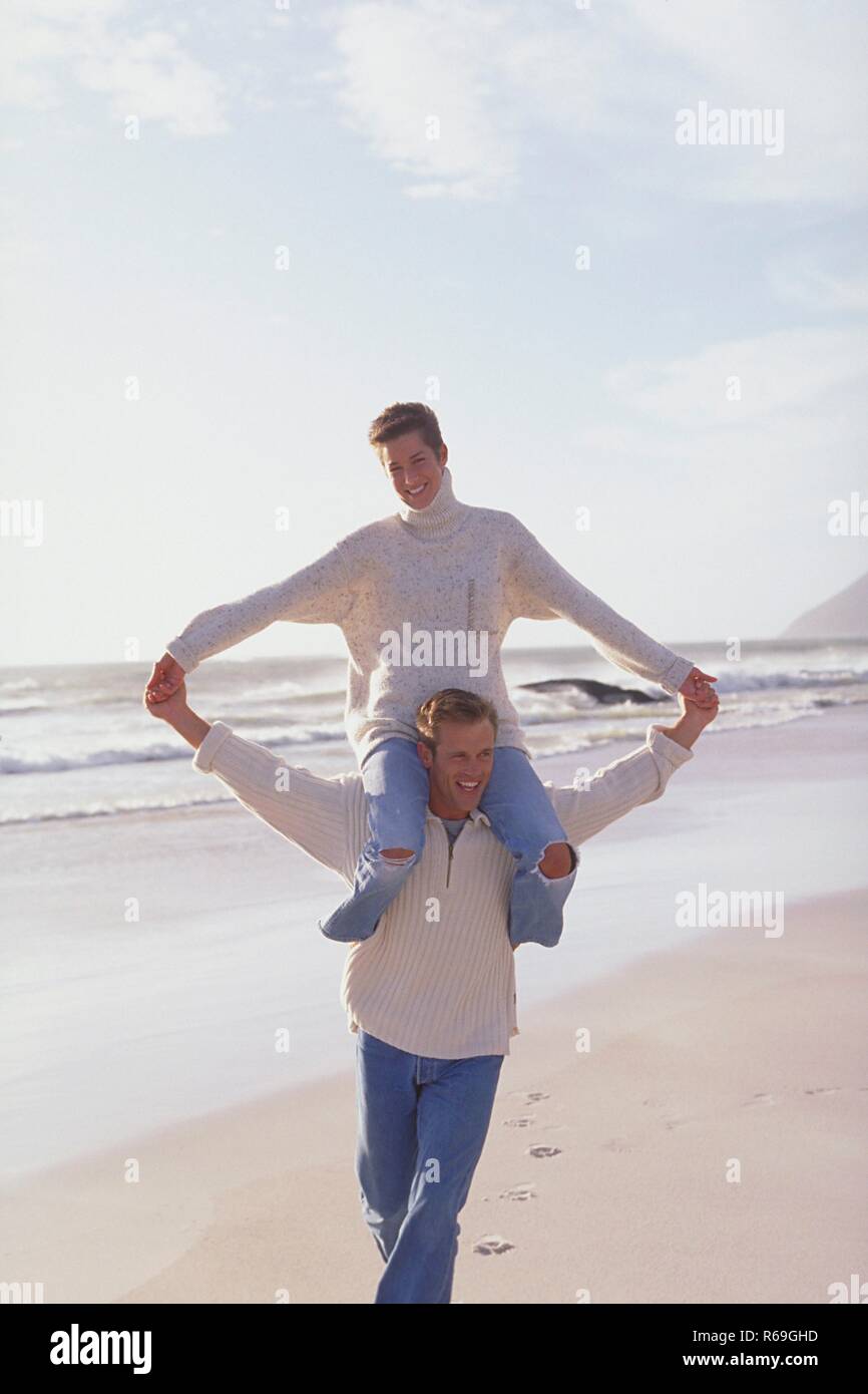 Strandszene, Mann laeuft mit seiner auf den Schultern sitzenden Frau am Strand entlang, beide bekleidet mit naturfarbenen Wollpullovern und Jeans Stock Photo