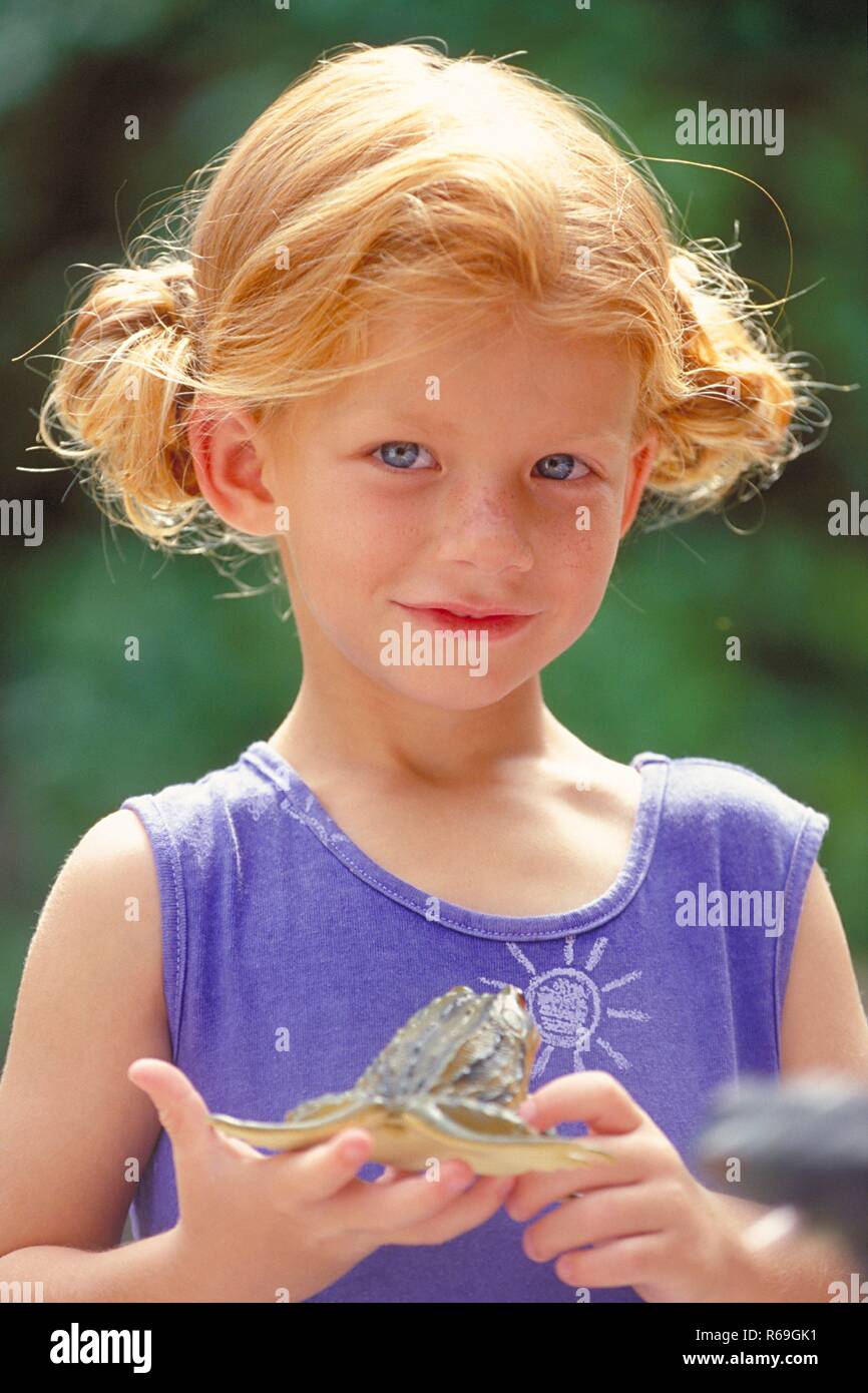 Portrait, Outdoor, rotblondes sommersprossiges Maedchen mit blauen Augen, 5 Jahre alt, bekleidet mit blauem aermellosen Kleid, haelt einen Gummifrosch in ihren Haenden Stock Photo