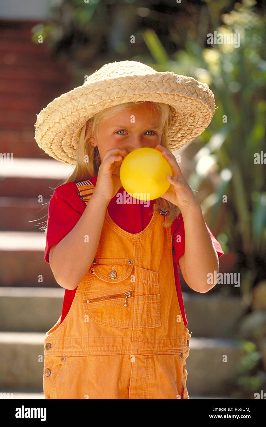 Portrait, Outdoor, Halbfigur, blondes Maedchen, 6 Jahre alt, bekleidet mit  rotem T-Shirt, oranger Latzhose und Strohhut blaest einen gelben Luftballon  auf Stock Photo - Alamy