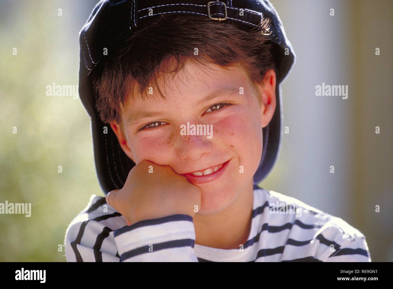 Portrait, Outdoor, Nahaufnahme, lustiger sommersprossiger Junge, 8 Jahre alt,  bekleidet mit blau-weiss gestreiftem T-Shirt und Baseballkappe mit Schild nach hinten Stock Photo