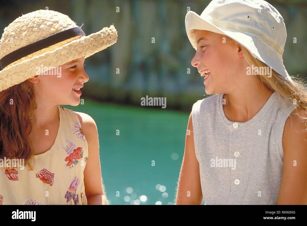 Outdoor, Nahaufnahme, Profil von zwei sich unterhaltenden lachenden Maedchen mit langen Haaren, 8-10 Jahre alt, bekleidet mit hellen Sommerkleidern und Hueten am Rand eines Schwimmbeckens Stock Photo