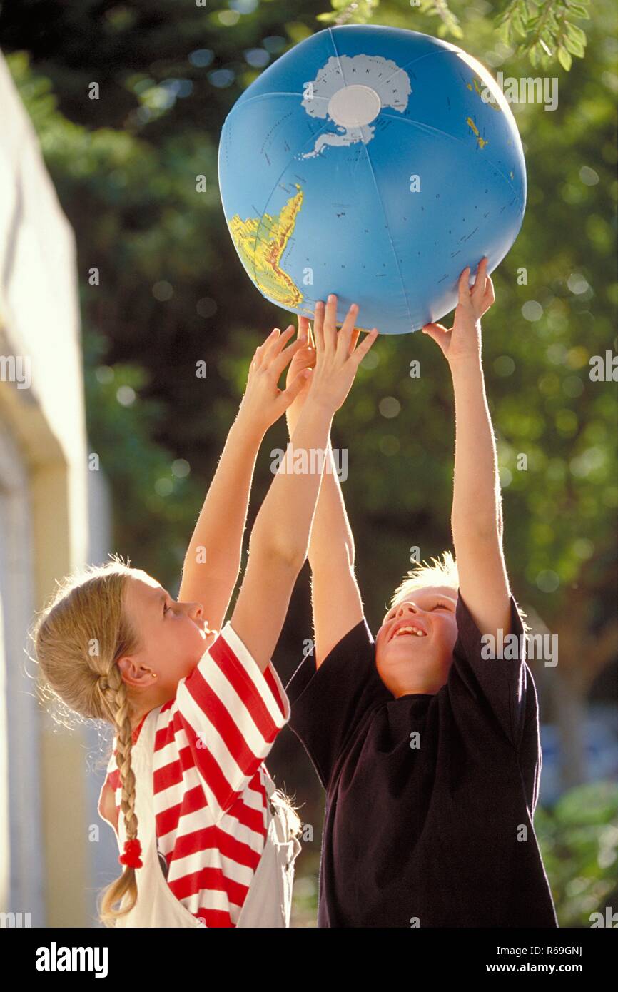 Outdoor, Halbfigur, blondes Maedchen mit Zoepfen und blonder Junge, 8 Jahre alt, spielen lachend mit einem Wasserball bedruckt mit der Erde Stock Photo