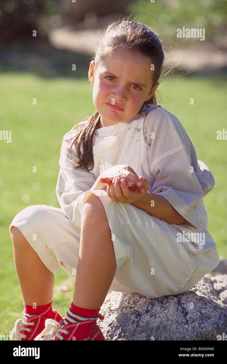 Portrait, Outdoor, Maedchen, 8 Jahre alt, mit langen braunen Haaren bekleidet mit heller Hose und roten Basketballschuhen sitzt traurig auf einem Stein im Park Stock Photo