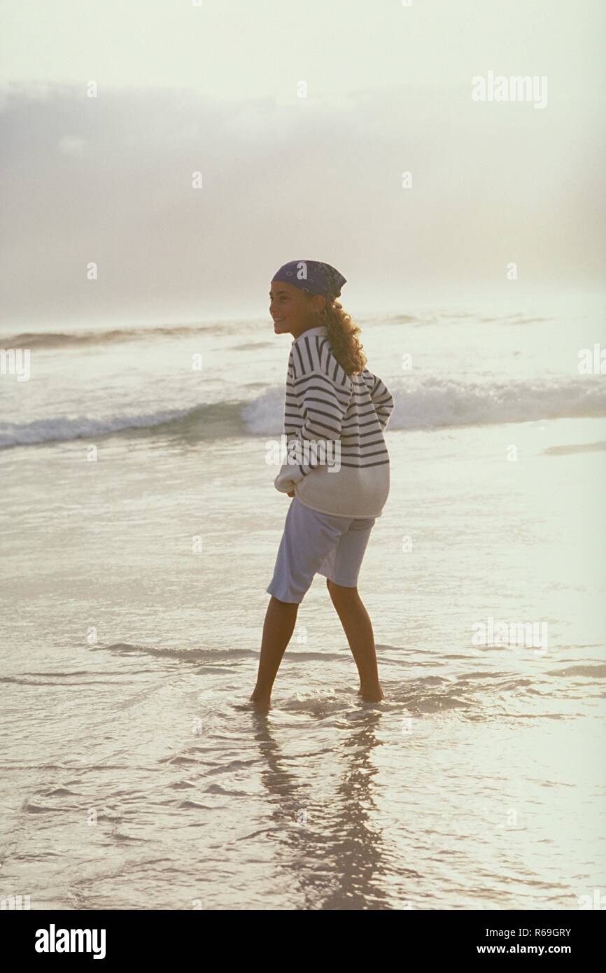 Strandszene, barfuessiges 10 Jahre altes Maedchen mit lockigem braunen Haar bekleidet mit Kopftuch, weisser Hose und geringeltem  Pullover steht lachend im seichten Wasser Stock Photo